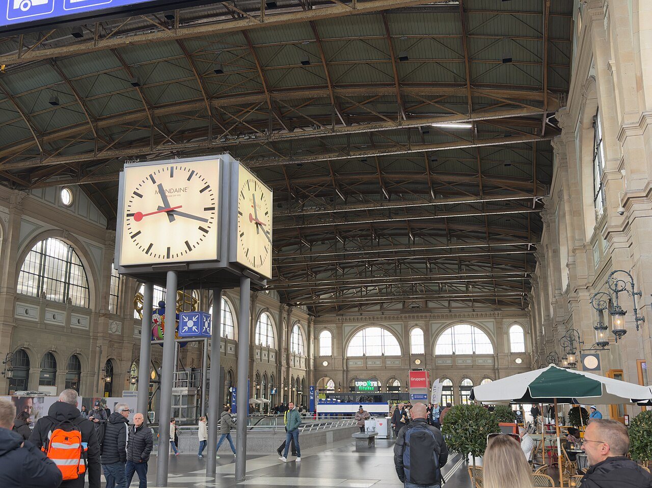 A large four-sided Mondaine clock on posts in the main hall of Zurich Hauptbahnhof, with travelers walking beneath the station's iron roof