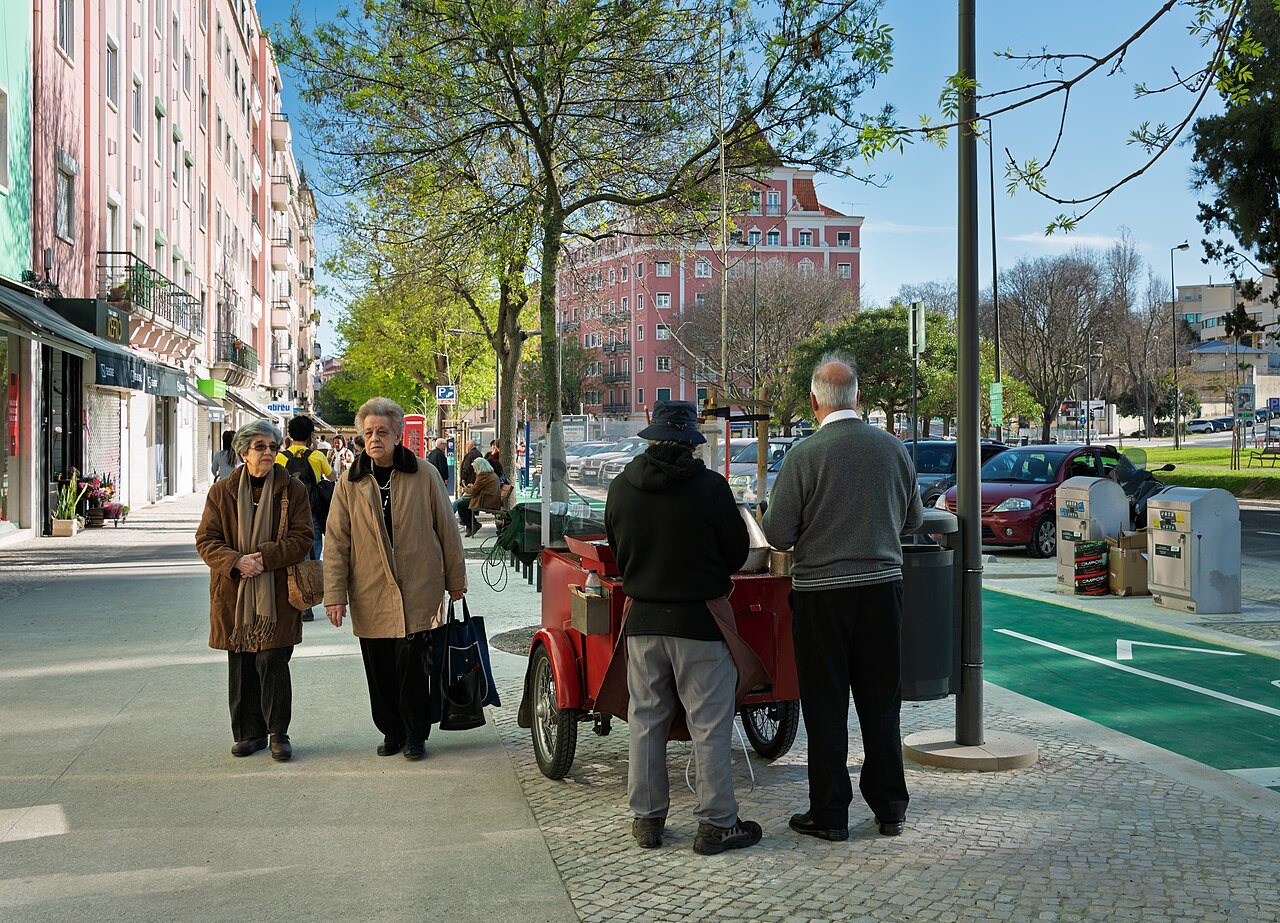 A roasted chestnut vendor in a black hat beside his red cart on a tree-lined street at Praça de Londres in Lisbon