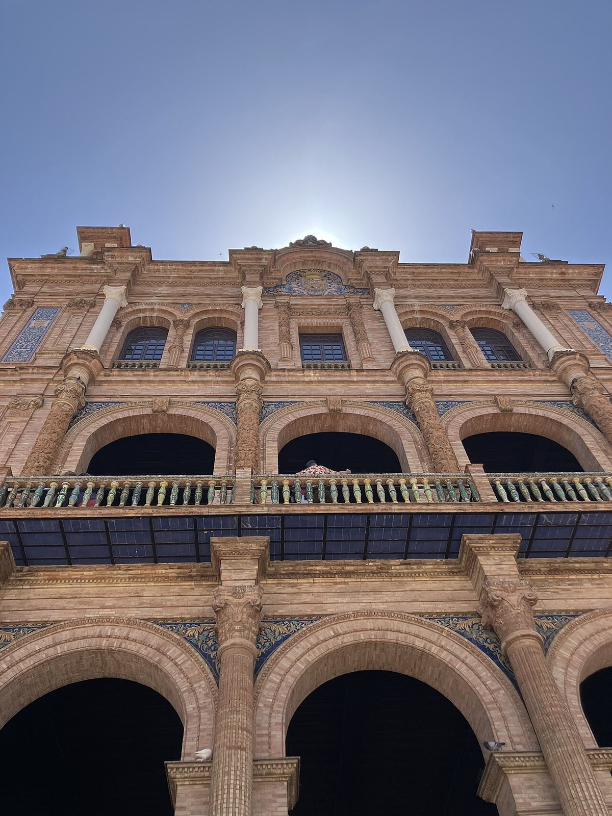 Looking up at a brick facade of the Plaza de España in Seville, with arched balconies, ceramic balustrades and blue tile panels against a clear sky