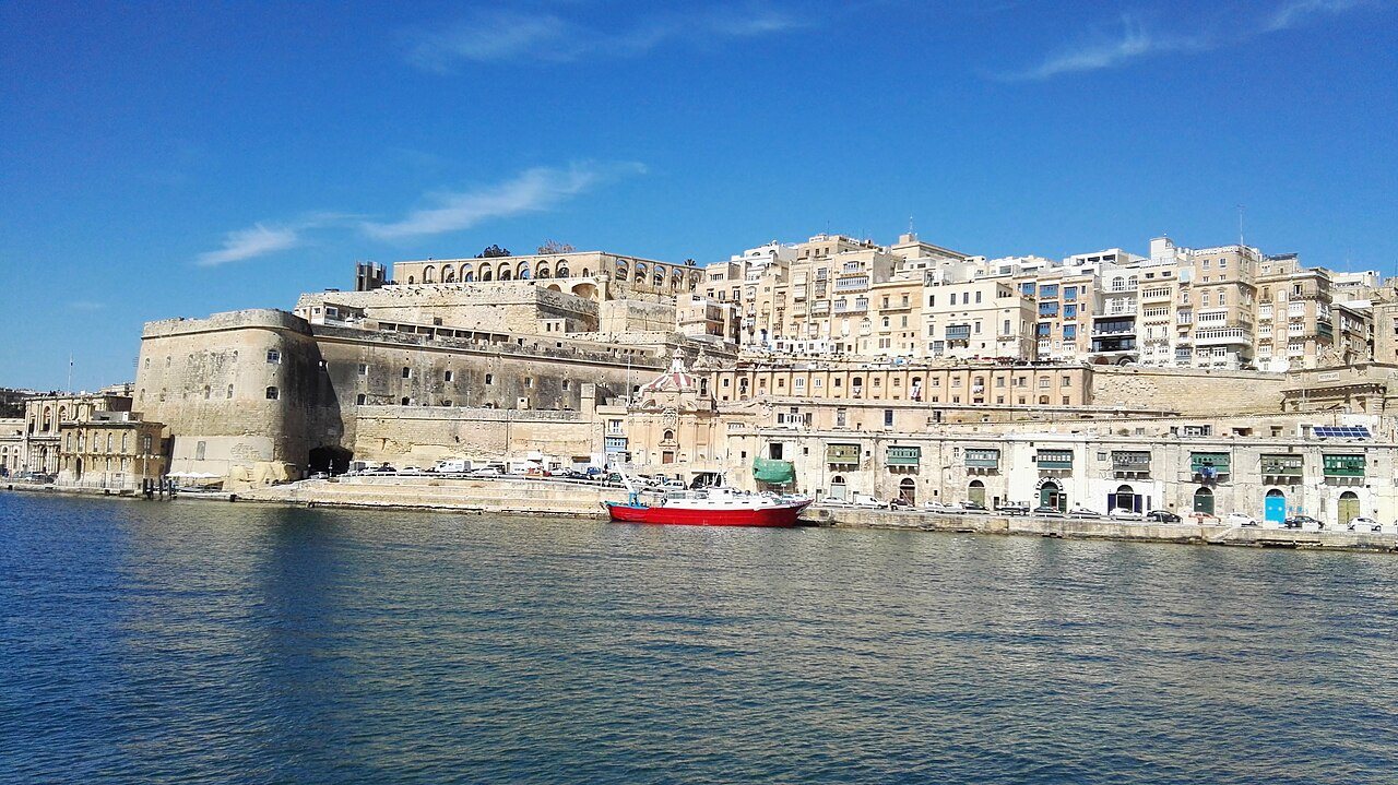 Valletta's honey-colored stone bastions and buildings rising above the Grand Harbour, with the arches of the Upper Barrakka Gardens at the top and a red boat moored on the waterfront